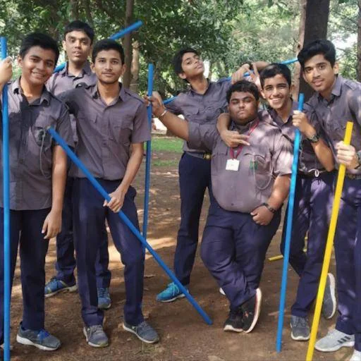 A group of boys in grey shirts and navy trousers holding blue and yellow poles in a wooded area during a school camp.