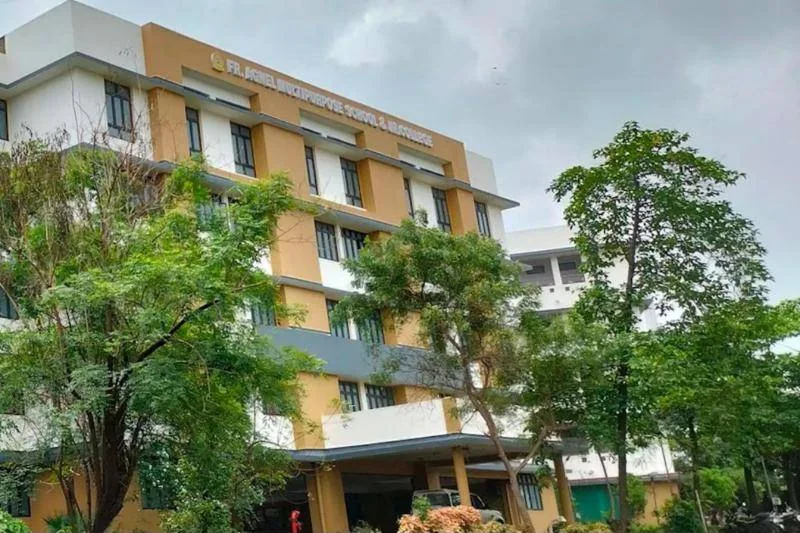 The yellow and white school building seen from the grounds, with tall trees in front and a grey monsoon sky above.